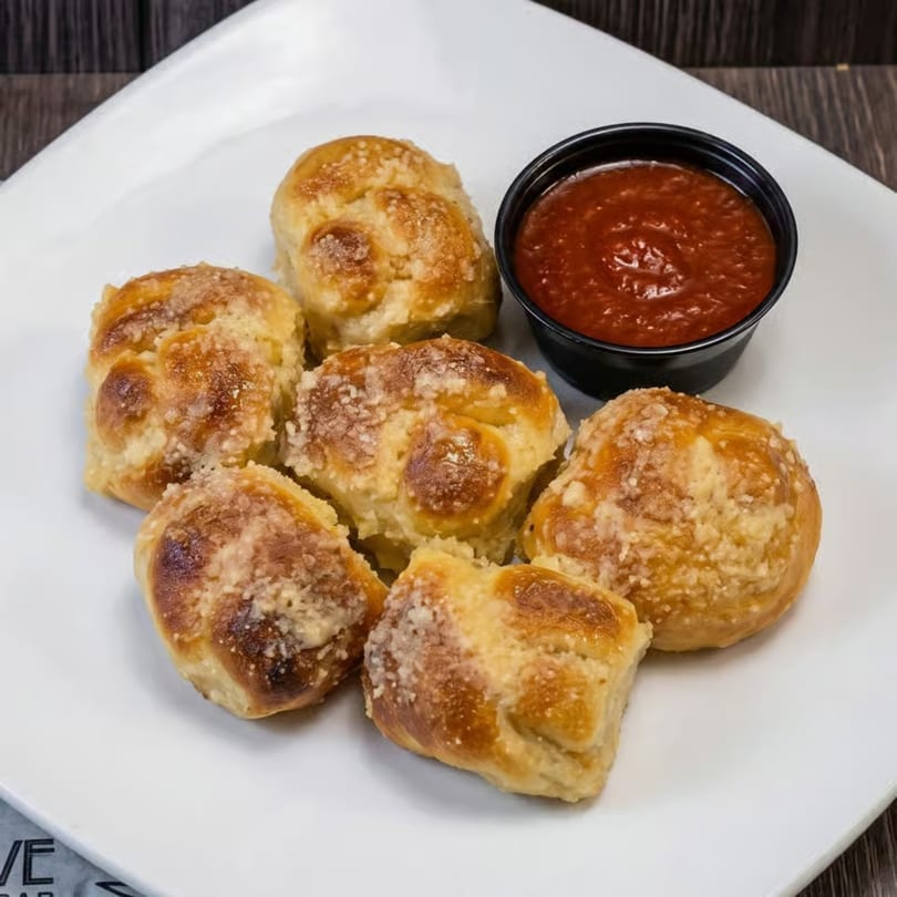 Six golden-brown garlic knots dusted with Parmesan cheese, served with a side of marinara sauce.